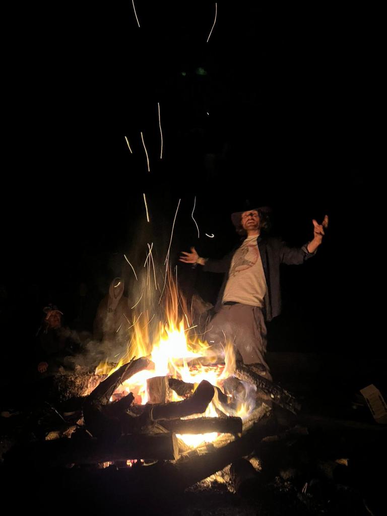 A storyteller stands beyond a blazing campfire, arms wide in the middle of a story, against a dark nightime background.