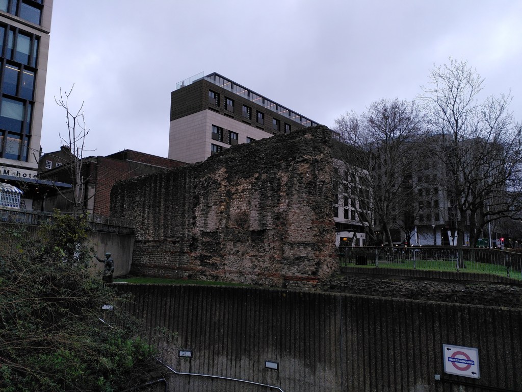 The Roman wall above a modern concrete underpass, with an office block in the background.